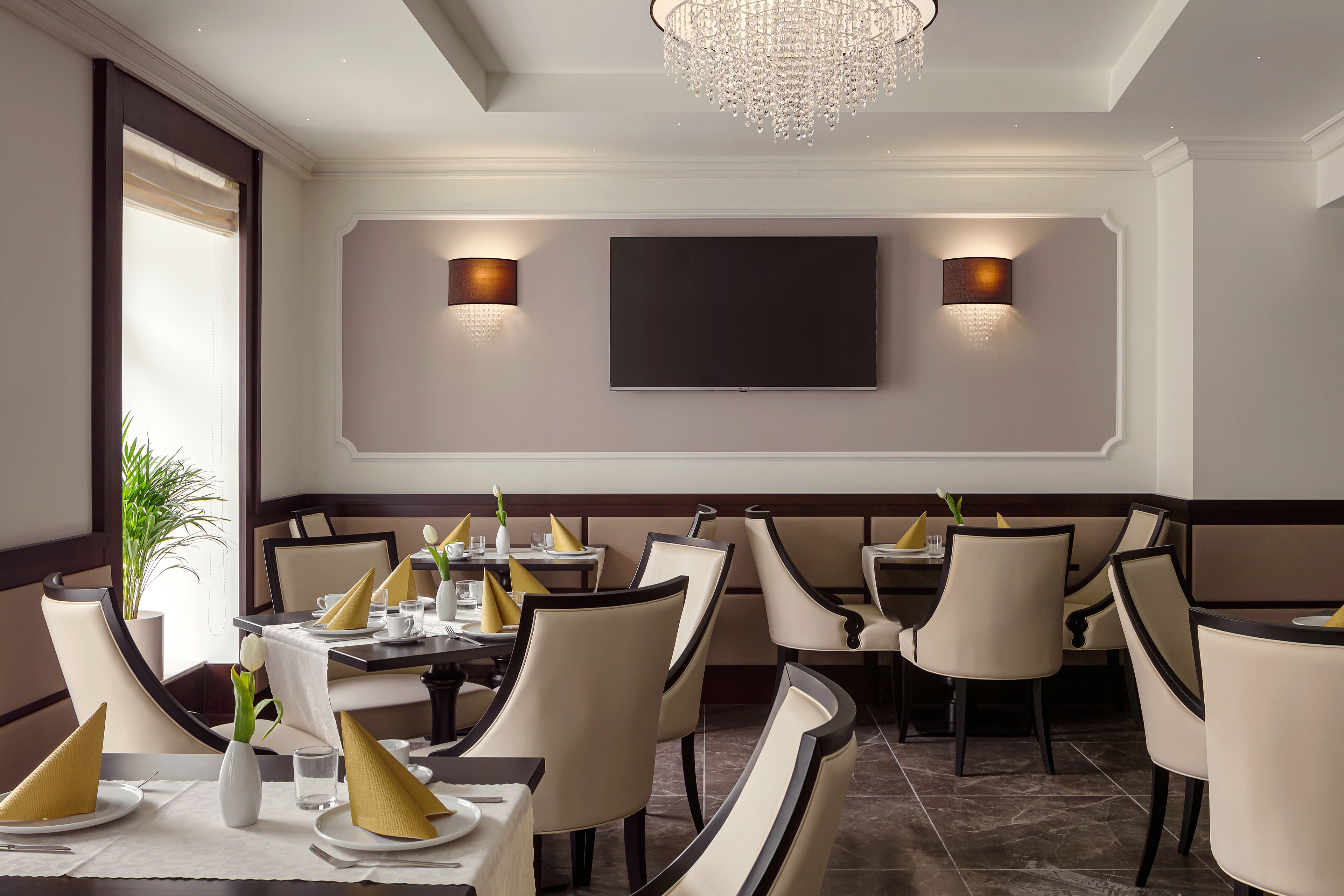 A restaurant with a modern interior, featuring round tables, beige chairs, and a large flat-screen TV mounted on the wall.