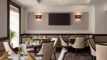 A restaurant with a modern interior, featuring round tables, beige chairs, and a large flat-screen TV mounted on the wall.