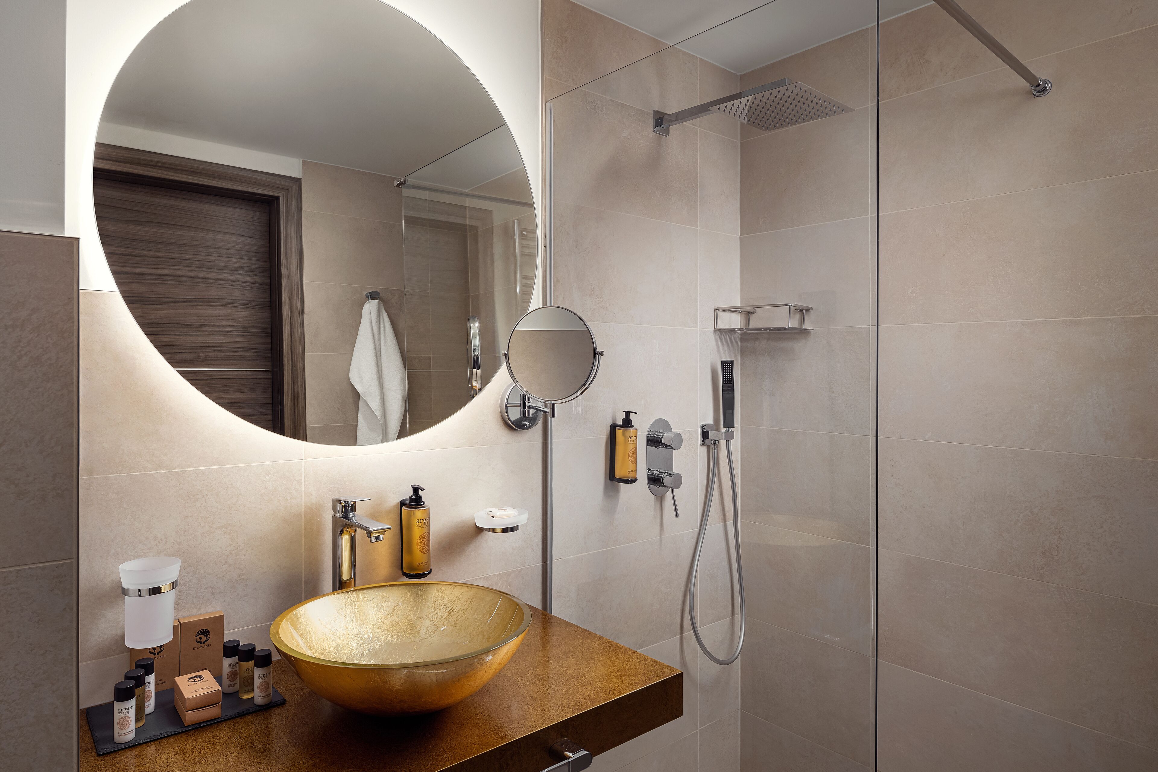 Une salle de bain moderne avec un grand miroir rond, un meuble-lavabo en bois, un lavabo en forme de bol en verre et une douche à l’italienne.