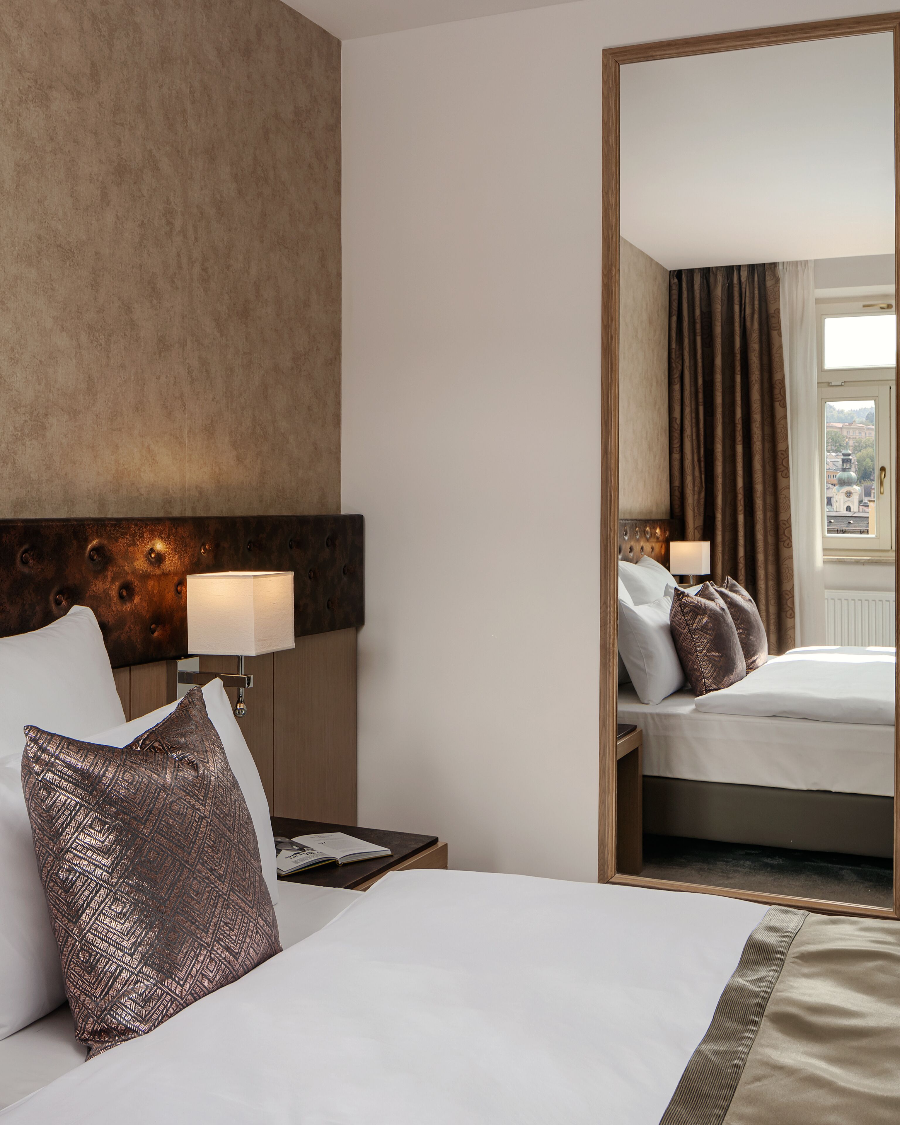 A hotel room with a large bed, a nightstand with a lamp, a headboard, and a view of the city through a window.