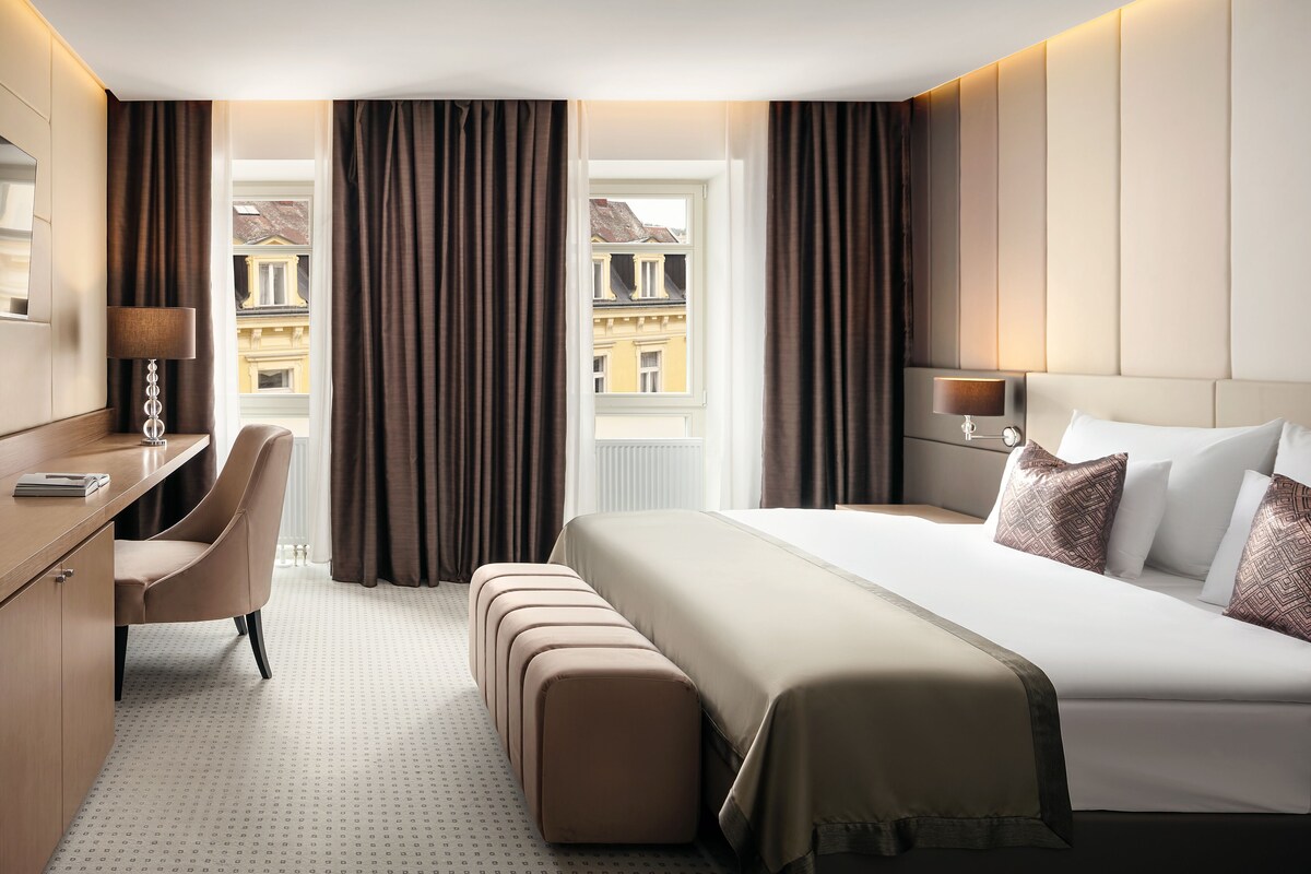 Une chambre d’hôtel avec un lit, un bureau, une chaise, un banc et une vue sur les immeubles par la fenêtre.