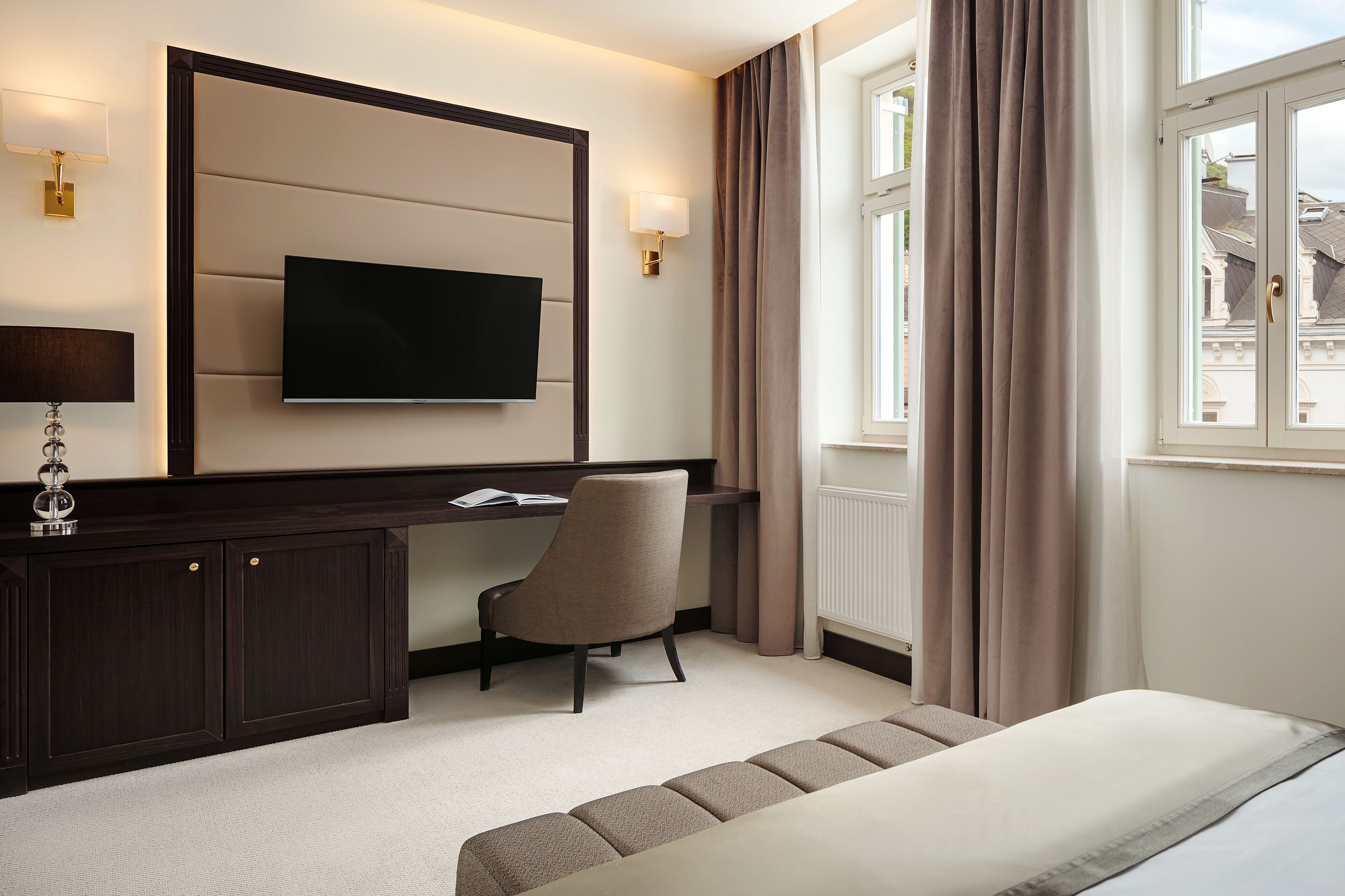 Habitación de hotel con una televisión de pantalla plana grande montada en la pared, un escritorio con una silla, una cama con cabecera y una ventana con cortinas.
