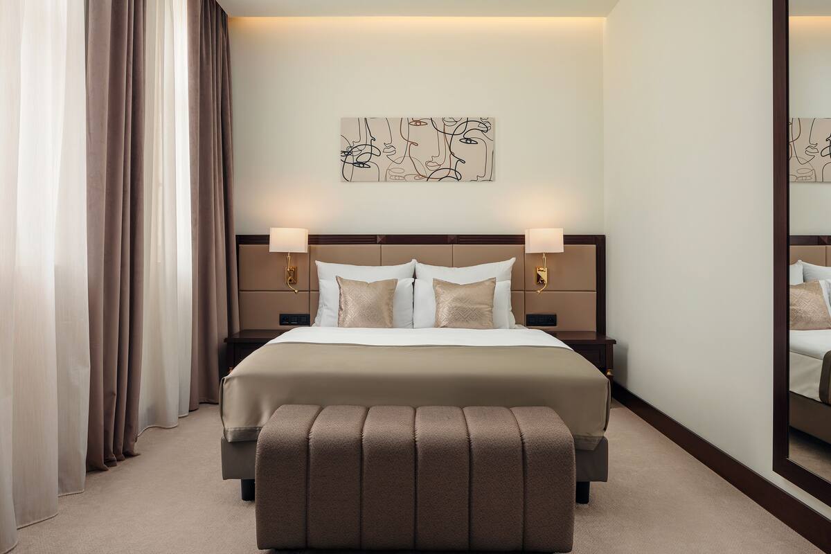 Una habitación de hotel con una cama grande, dos mesitas de noche con lámparas, un cabecero, un espejo amplio y una obra de arte en la pared.