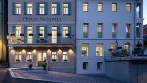 Een hotel met twee verdiepingen, voorzien van een balkon op de tweede etage, een bord met daarop 'HOTEL SAXONIA', en een geplaveid straatje ervoor.