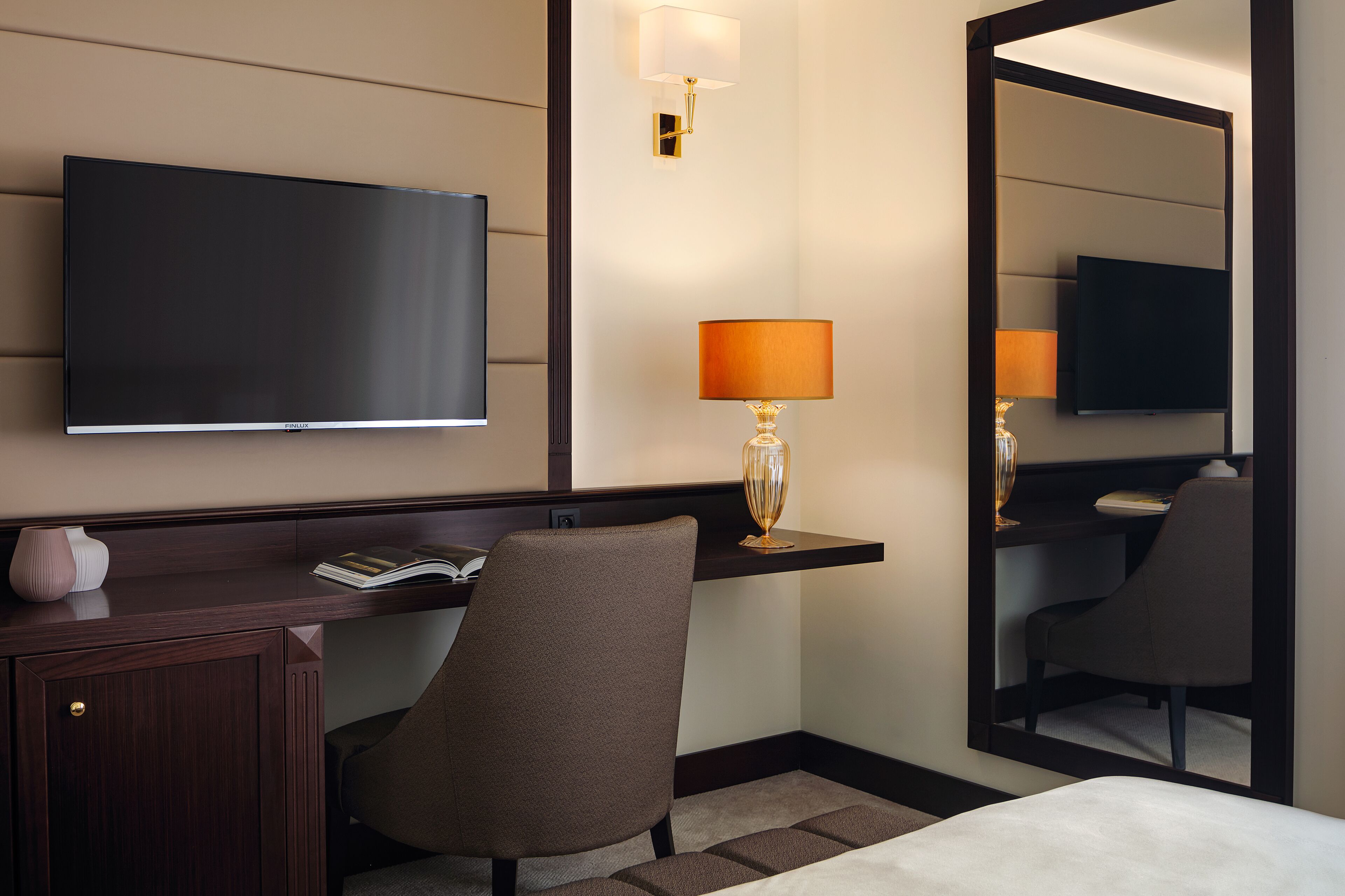 A hotel room with a flat-screen TV, a desk with a lamp, a chair, and a bed.