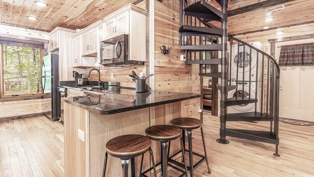 A modern kitchen with wooden cabinets, a central island with stools, a built-in oven, and a spiral staircase.