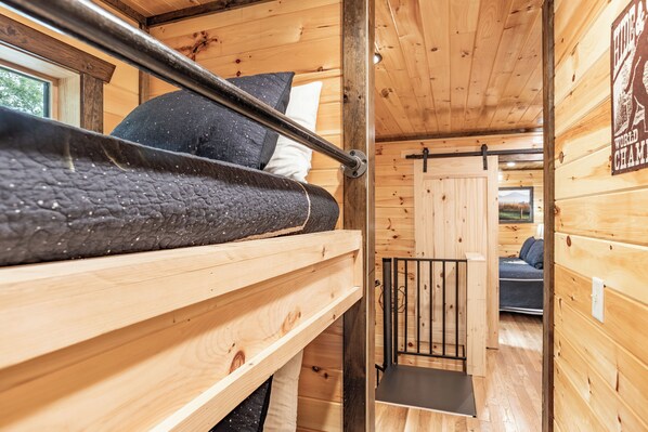 Interior de una cabaña de madera con una litera, una pequeña silla y una puerta que da a otra habitación.