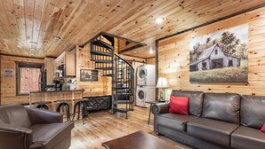 Un intérieur de chalet chaleureux, avec un revêtement mural en bois, un canapé, une table à manger et un coin cuisine doté d’un escalier en colimaçon.