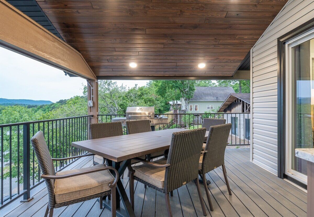 A covered outdoor deck with a wooden table and wicker chairs, overlooking a residential area.