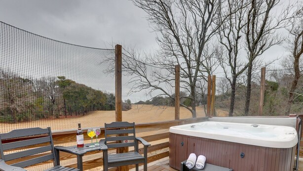 An outdoor deck with a hot tub, two wooden chairs, a small table with a bottle and glasses, and a netted enclosure. - Luxury Golf Villa w/ Hot Tub  Stunning Golf Course Views Near OBX Beaches (Kitty Hawk)