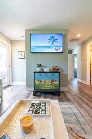 A living room with a flat-screen TV mounted on the wall, a wooden coffee table with a candle, and a decorative dresser with painted artwork. - Luxury Golf Villa w/ Hot Tub Stunning Golf Course Views Near OBX Beaches (Kitty Hawk)