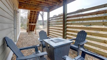 Une terrasse extérieure avec une table et quatre chaises Adirondack, une pergola en bois au-dessus et une vue sur un espace clos planté d’arbres.