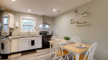 Une cuisine moderne avec des meubles blancs, une table à manger en bois et des chaises blanches. On y trouve des étagères intégrées ornées d’objets décoratifs et une fenêtre au-dessus de l’évier.
