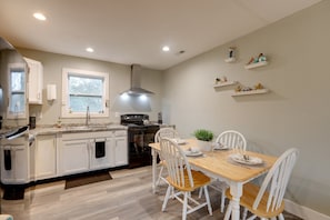 A modern kitchen with white cabinetry, a wooden dining table, and white chairs. There are built-in shelves with decorative items and a window above the sink. - Luxury Golf Villa w/ Hot Tub Stunning Golf Course Views Near OBX Beaches (Kitty Hawk)
