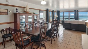 A dining area with a large wooden table and chairs, a cabinet with glass doors, and a view of the sea through large windows. - Griners Life Saving$ Station on Lake Michigan w/ Private Beach, Pet-Friendly (Frankfort)
