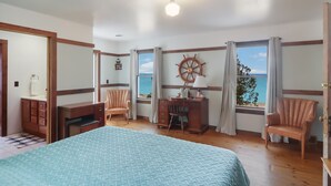 A bedroom with a bed, desk, chairs, a ship's wheel decoration, and a view of the sea through large windows. - Griners Life Saving$ Station on Lake Michigan w/ Private Beach, Pet-Friendly (Frankfort)