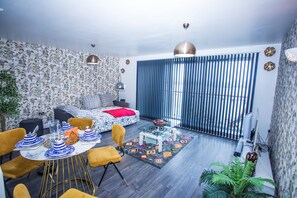 A modern living room with a sofa, dining table, and chairs. There is a large window with blinds, a ceiling light, and a plant. - Remarkable 2-bed Apartment With Sea View (London)