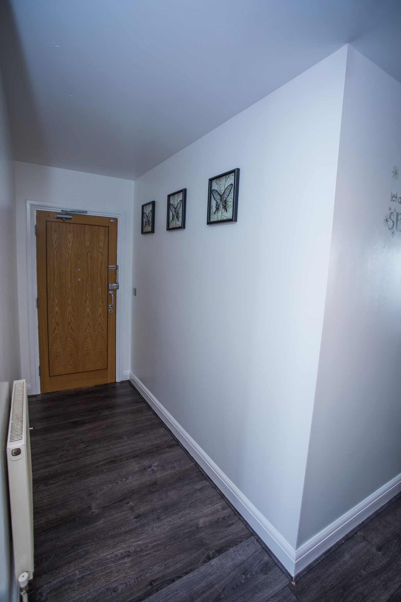Un couloir avec une porte en bois, des murs blancs et un plancher en bois foncé. Trois tableaux encadrés sont accrochés au mur.