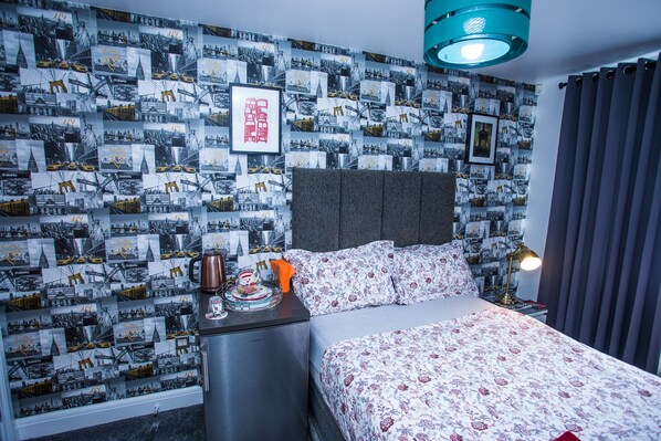 A hotel room with a patterned wallpaper, a bed with floral bedding, a nightstand with a lamp, and a coffee maker. - Remarkable 2-bed Apartment With Sea View (London)