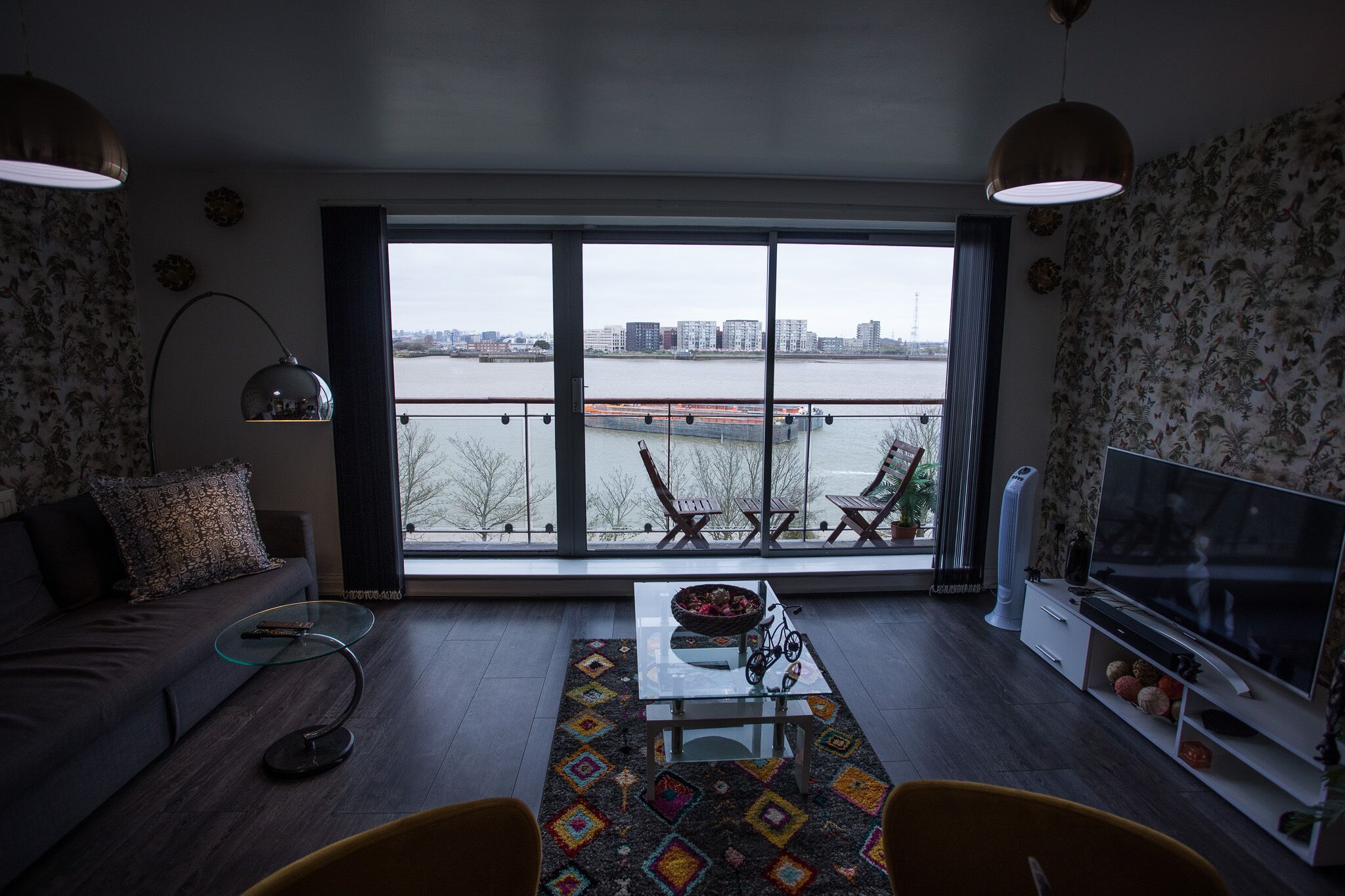 A modern living room with a large window, a sofa, a coffee table, a TV, and a balcony with outdoor furniture.