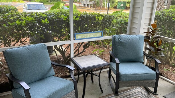 A porch with two blue cushioned chairs and a small table, a 'Welcome to the BEACH HOUSE' sign, and a view of the street and greenery. - Beautifully Decorated Condo with Pool and Fitness Center! Pet Friendly! (Saint Simons Island)