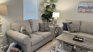 A living room with two grey sofas, patterned cushions, a wooden coffee table, a potted plant, a lamp, and a painting on the wall. - Beautifully Decorated Condo with Pool and Fitness Center! Pet Friendly! (Saint Simons Island)