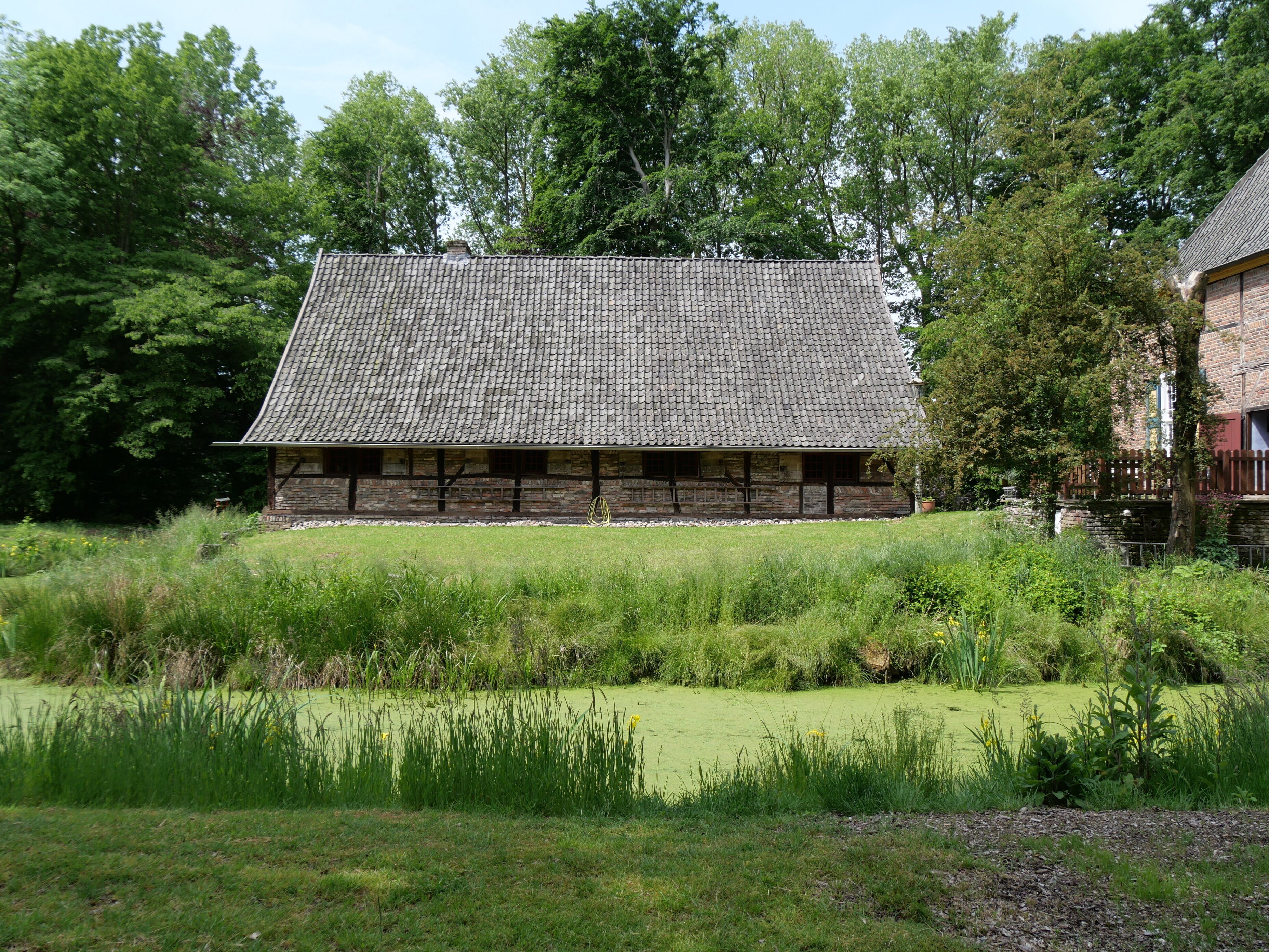 A traditional stone building with a tiled roof, surrounded by lush greenery and a pond.