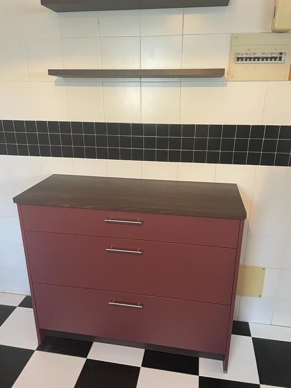 Een donkerrode kast met drie lades en een aanrecht, tegen een betegelde muur met een zwart-wit geruite vloer.