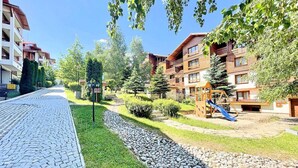 A residential area with a paved walkway, a playground with a slide, and multi-story buildings with balconies. - Luxury Apartments St Ivan Rilski SPA 4 (Bansko)