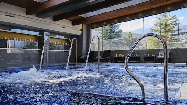 An indoor hot tub with metal railings, a view of mountains through large windows, and a modern interior design with wooden beams. - Luxury Apartments St Ivan Rilski SPA 4 (Bansko)