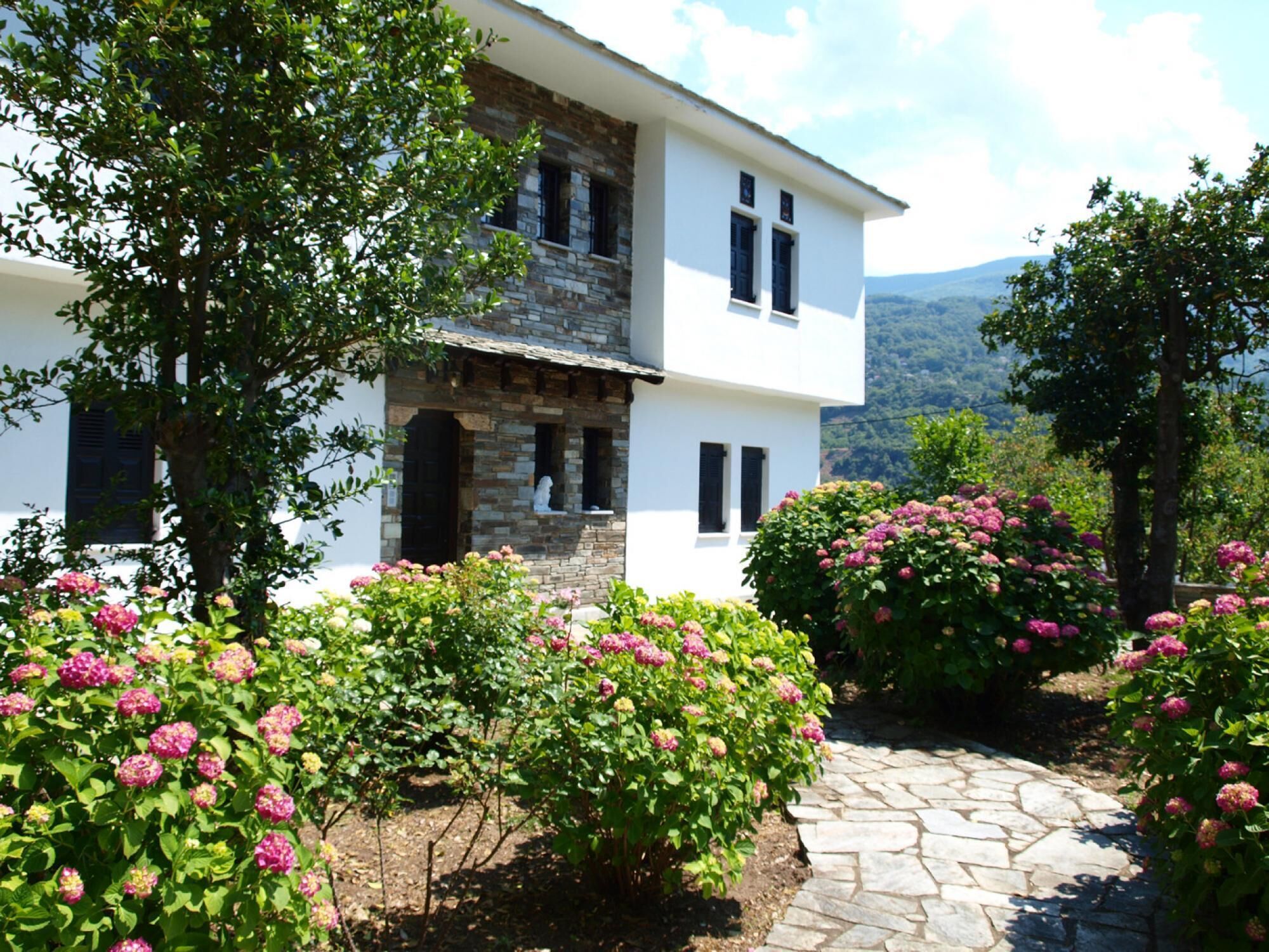 A two-story house with a stone facade, surrounded by lush greenery and flowering bushes.