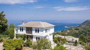 A white two-story house with a tiled roof, surrounded by greenery and overlooking a coastal landscape. - Villa Zina Studio 1 (Zagora-Mouresi)