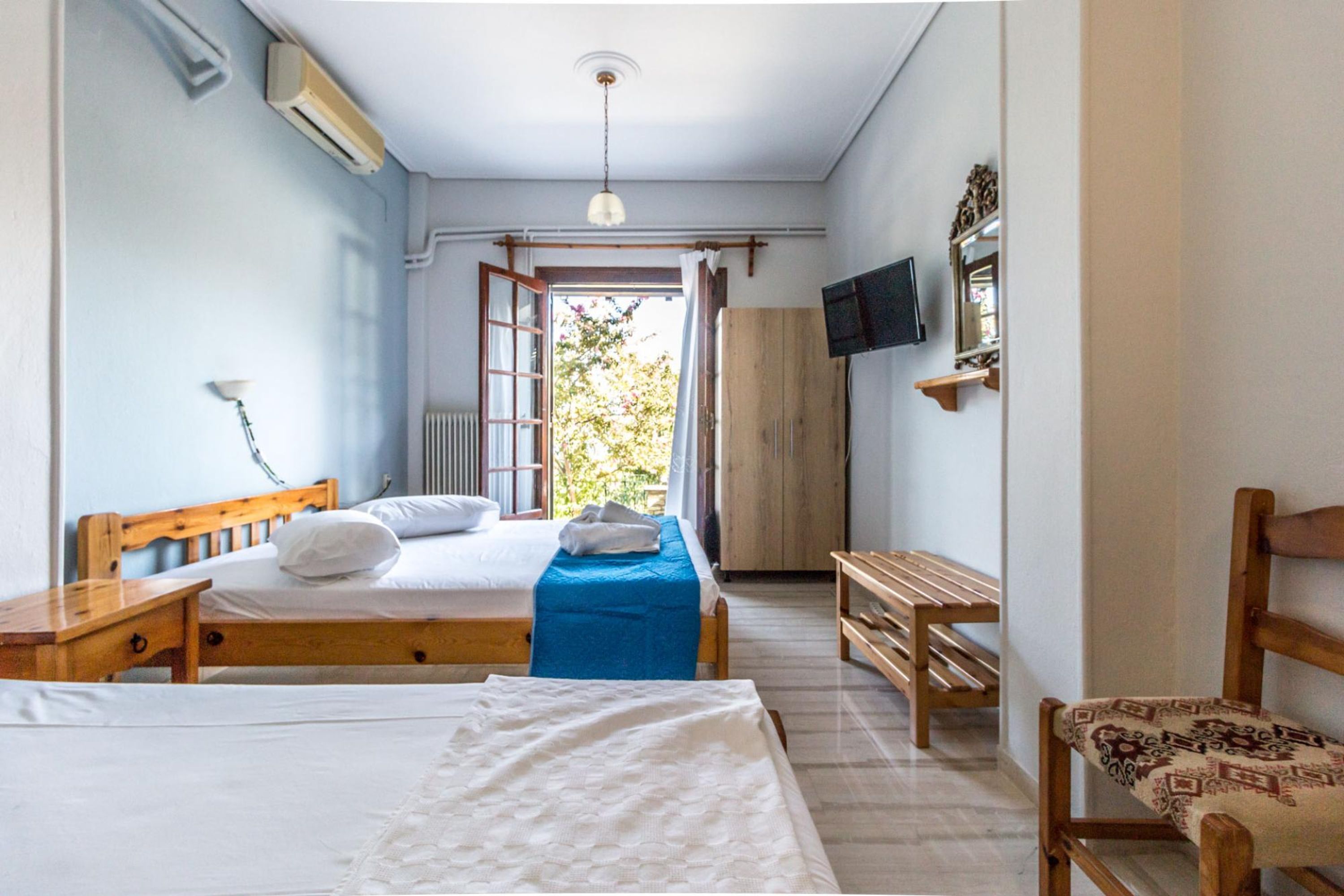 A bedroom with two beds, a wooden bench, a chair, a TV, and an open window.