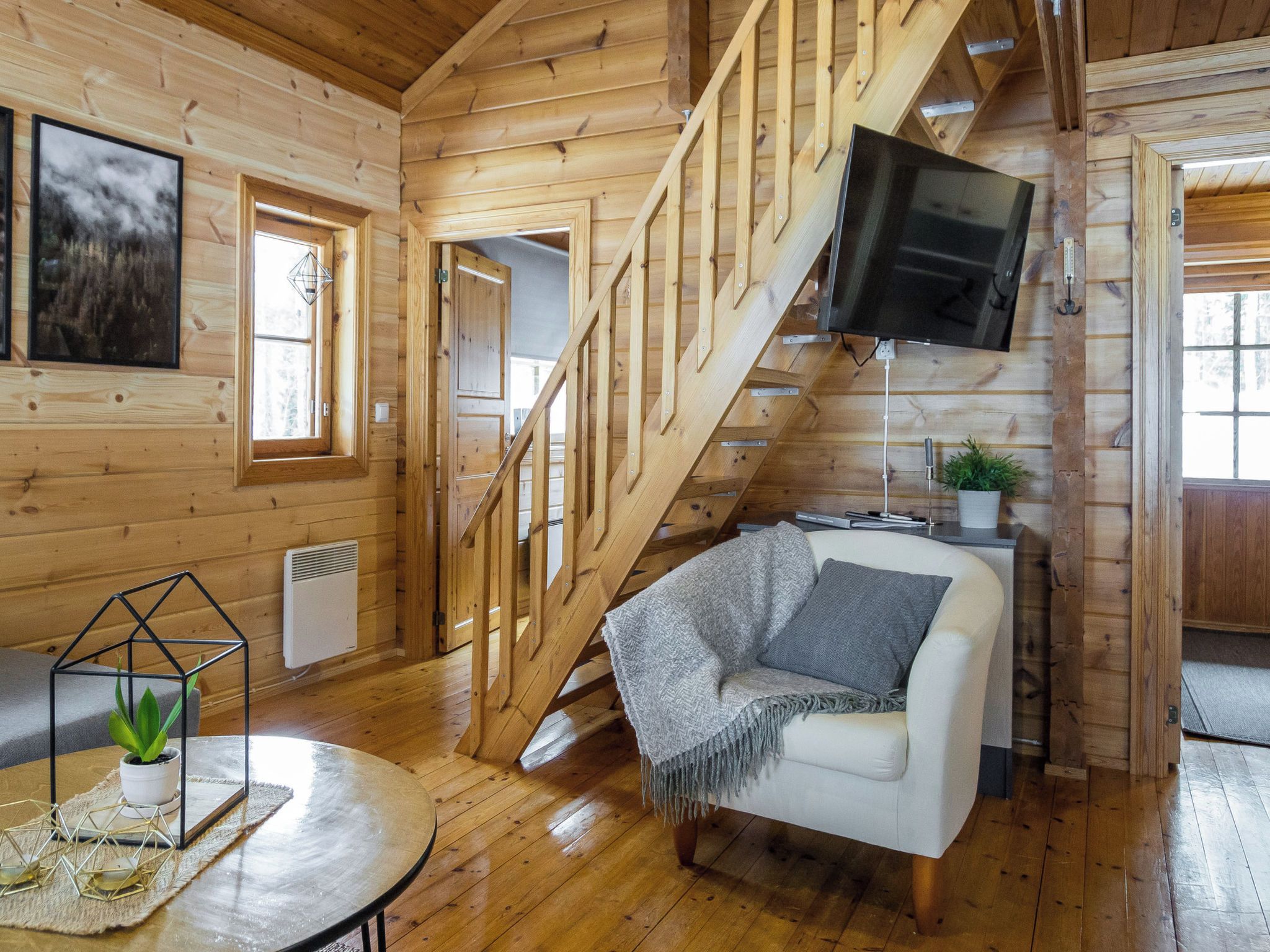 A wooden cabin interior with a staircase, a white sofa, a TV, and a small table with a plant.