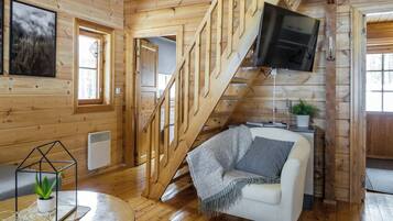 A wooden cabin interior with a staircase, a white sofa, a TV, and a small table with a plant.