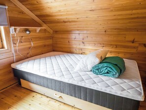 Una cama individual con colchón, almohadas y una manta verde en una habitación de madera con techo inclinado.