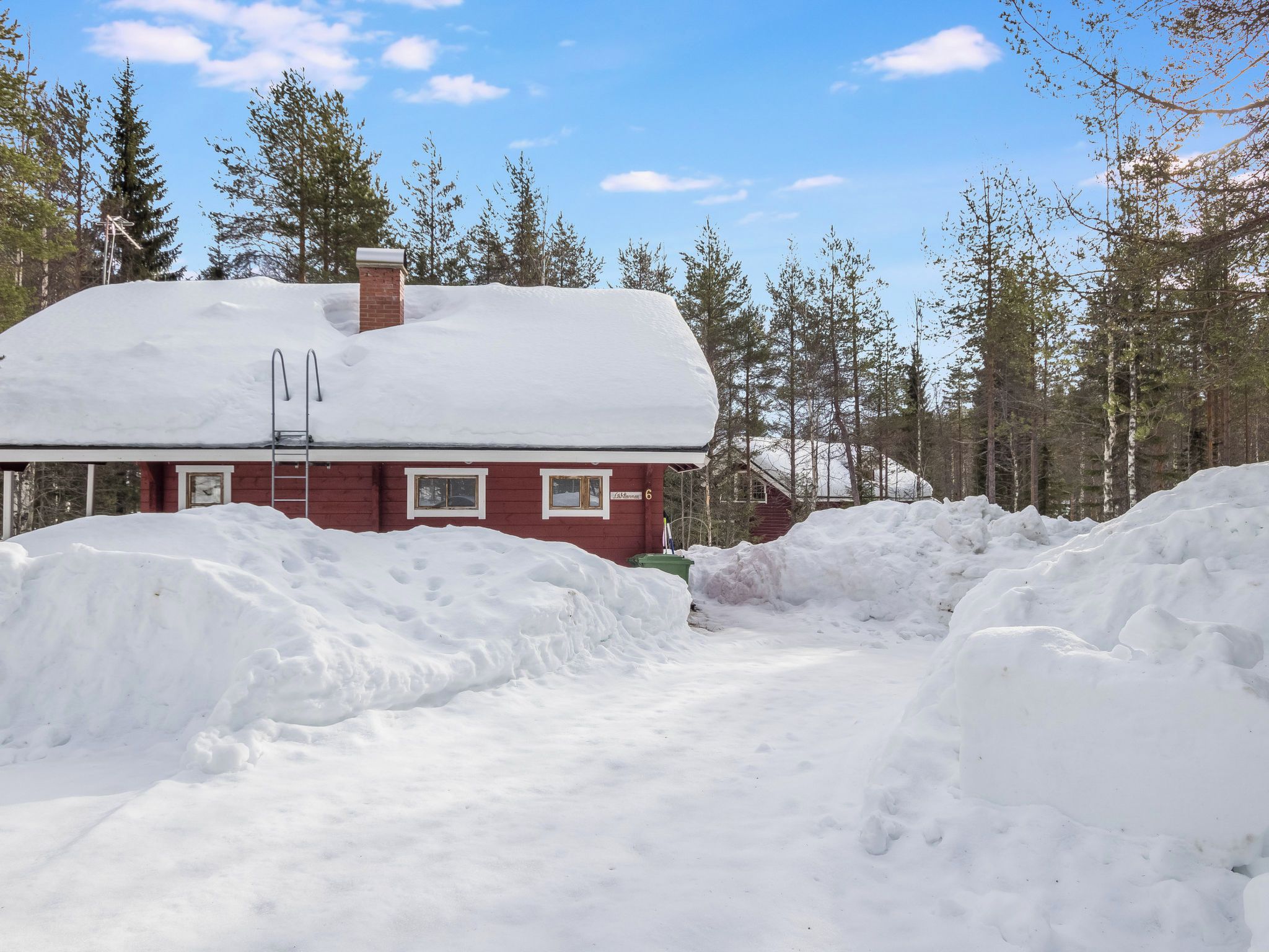 A red cabin with a snow-covered roof, surrounded by a forest and large snowbanks.