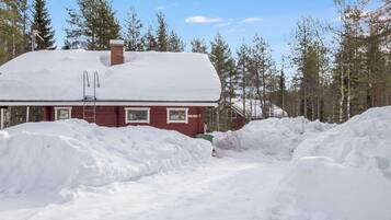 A red cabin with a snow-covered roof, surrounded by a forest and large snowbanks.