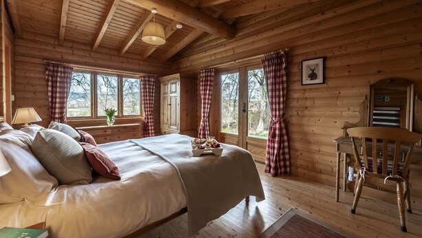 A wooden cabin bedroom with a bed, a chair, and a window with curtains. - Croft Kincardine -  a lodge that sleeps 8 guests  in 4 bedrooms (Boat of Garten)