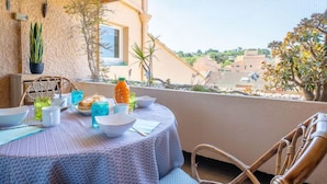 Un balcon avec une table dressée pour le dîner, agrémenté d’une nappe à motifs, de chaises, ainsi que de différents plats et boissons.