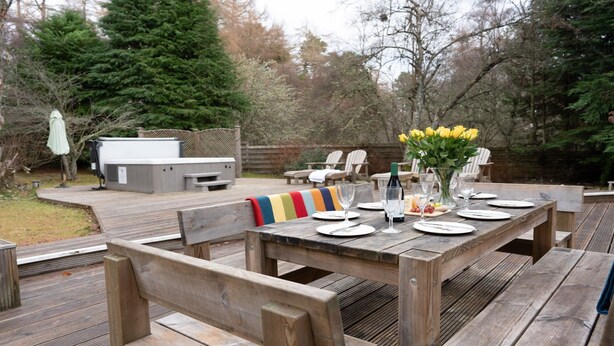 A wooden deck with a table set for dining, surrounded by chairs and a barbecue grill. - Four Pines Lodge -  a house that sleeps 8 guests  in 4 bedrooms (Dulnain Bridge)