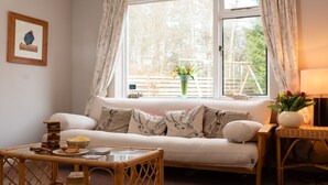 A cozy living room with a sofa, cushions, a small table with snacks, and a window with curtains. - Four Pines Lodge -  a house that sleeps 8 guests  in 4 bedrooms (Dulnain Bridge)