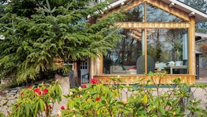 A modern wooden cabin with a glass enclosure, surrounded by lush greenery and flowering plants. - River House Garden Rooms -  a house that sleeps 2 guests  in 1 bedroom (Aviemore)