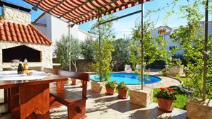 Une terrasse extérieure avec une table et des chaises en bois, un four en pierre, une piscine et des plantes en pot.