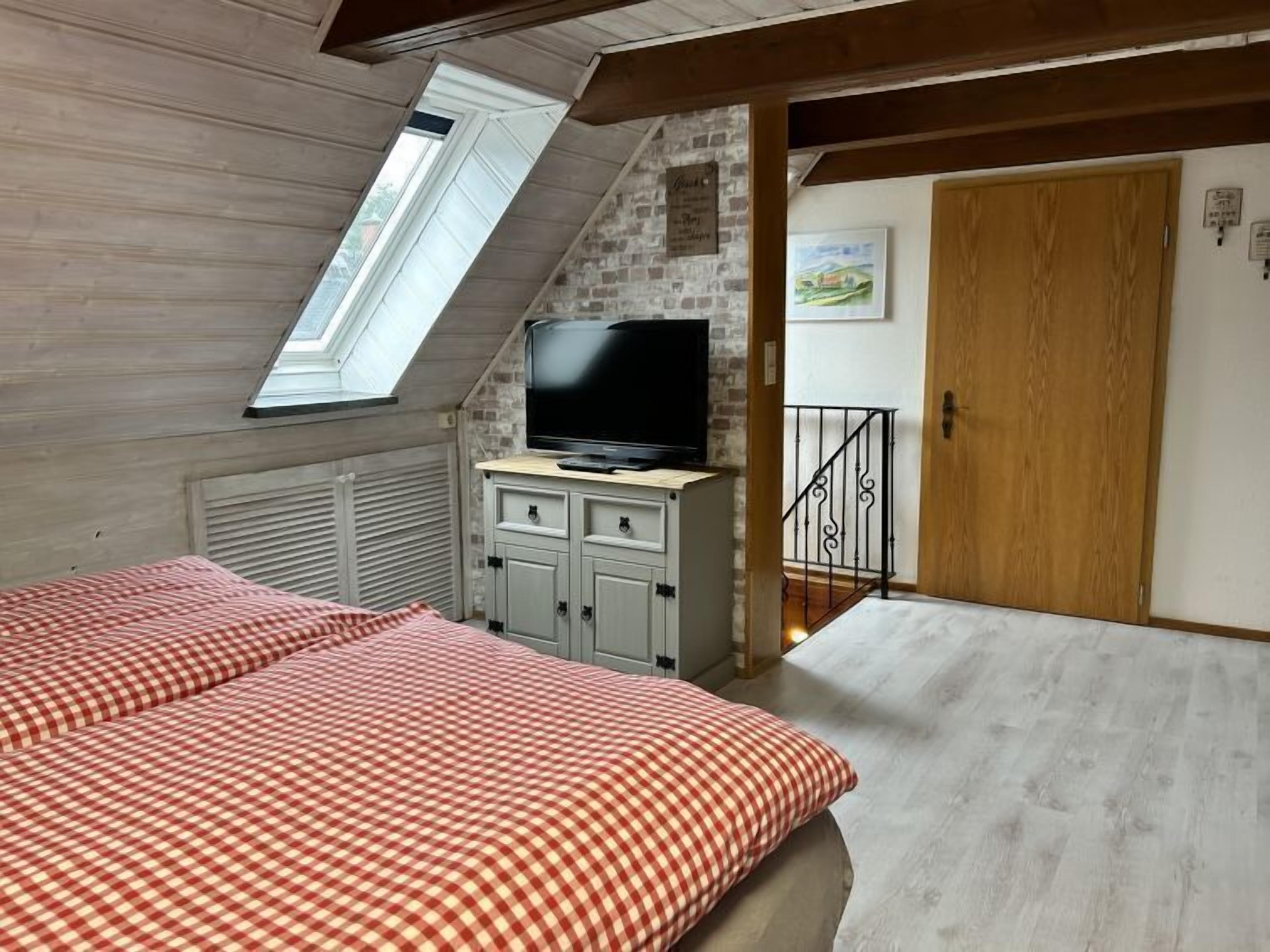 Une chambre avec un plafond en pente, un téléviseur posé sur une commode, une couvre-lit à carreaux et une porte en bois.