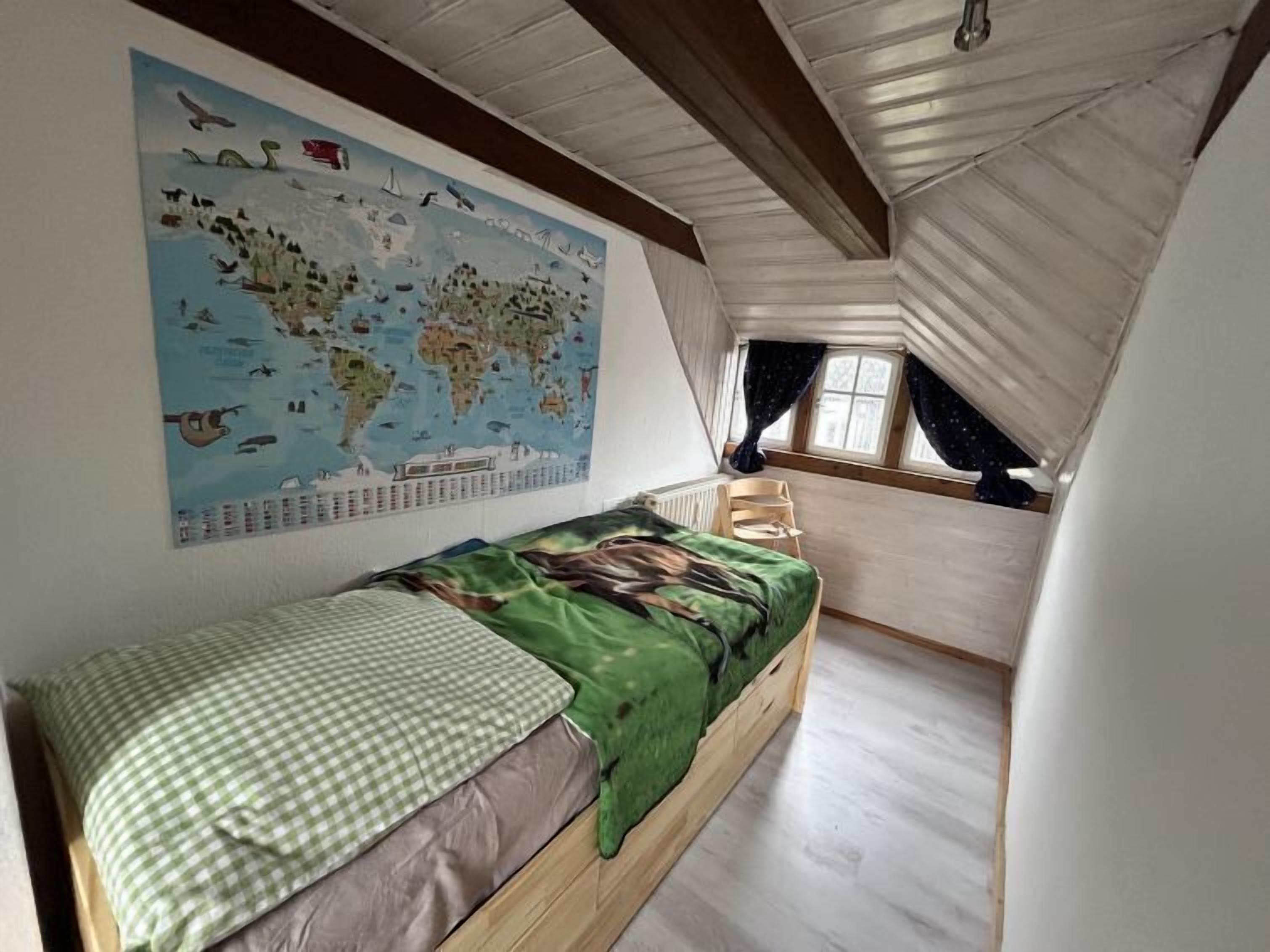 A small, cozy attic bedroom with a wooden bunk bed, a world map on the wall, and a window with curtains.