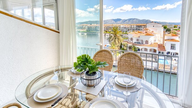 Un balcón con mesa de vidrio dispuesta para comer, con vista a un puerto deportivo y edificios.