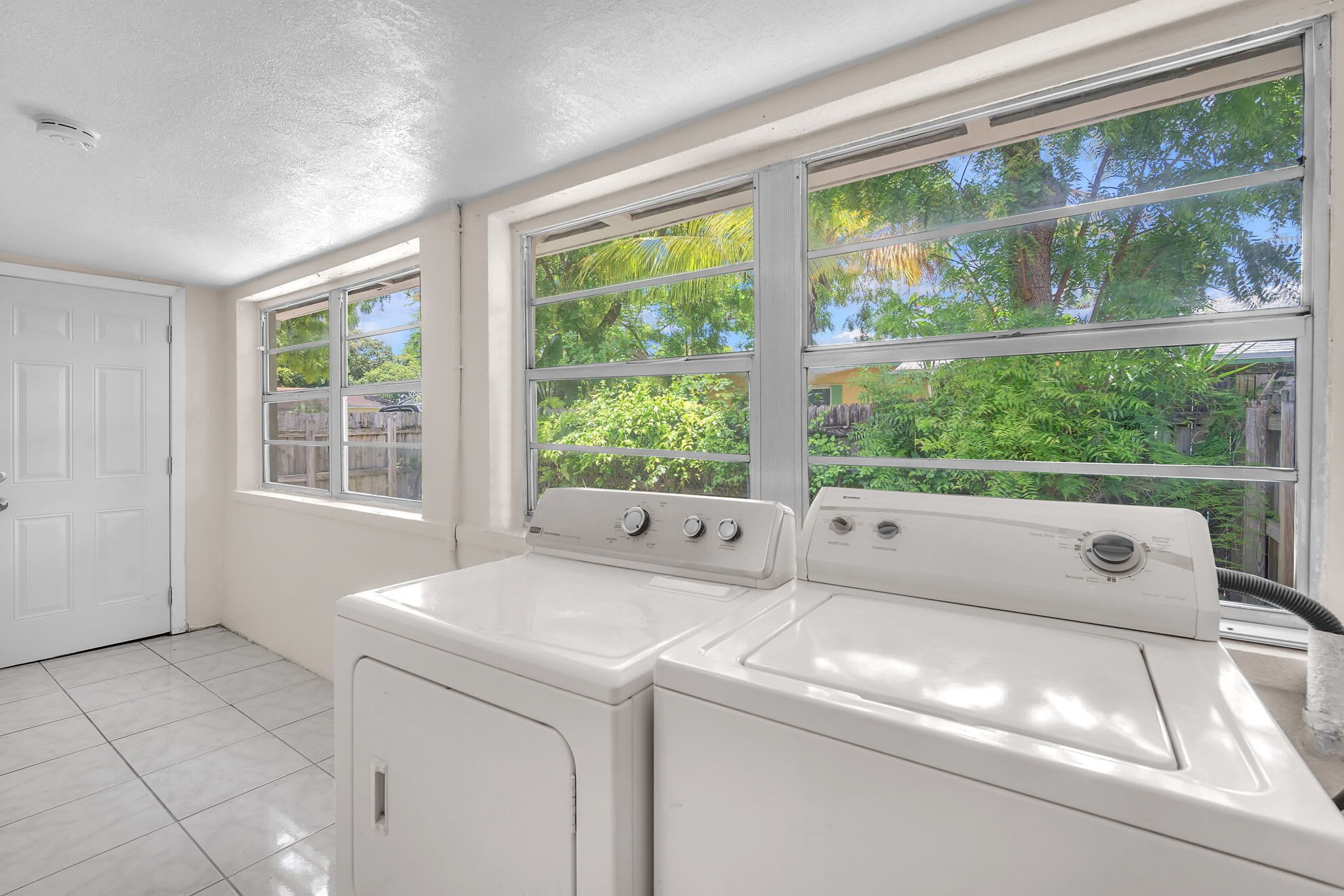 A laundry room with two white washers and dryers, a white door, and large windows with a view of greenery.