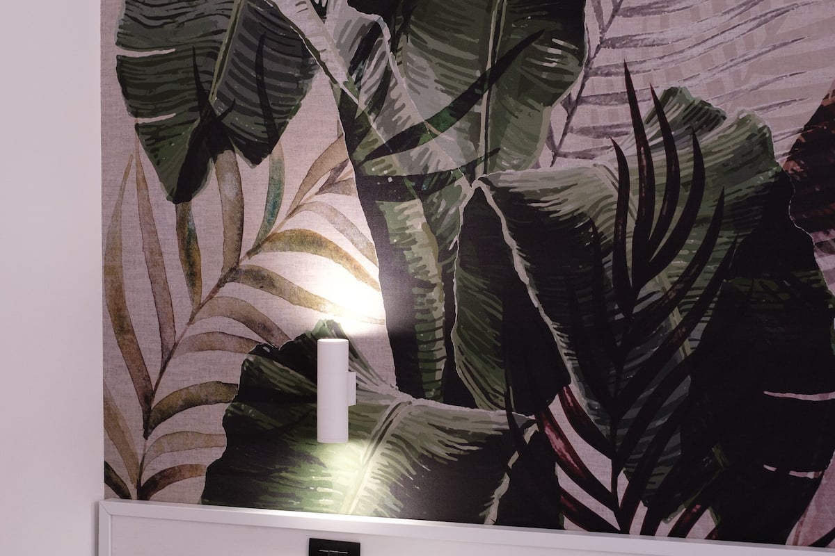 Une chambre d’hôtel avec un grand lit, du linge de lit blanc, une table de chevet, une lampe fixée au mur et un papier peint orné de motifs de feuilles tropicales.