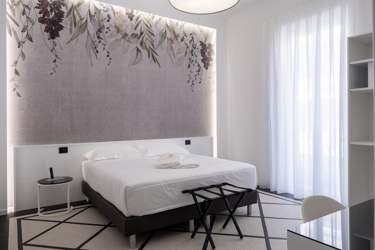 Une chambre d’hôtel moderne avec un grand lit, une table de chevet, une chaise et une vue sur l’extérieur à travers des rideaux transparents.